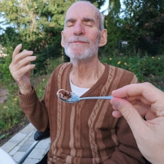 Älterer Mann sitzt im Rollstuhl im Freien, schließt genüsslich die Augen, während ihm jemand mit einem Löffel Essen anbietet.