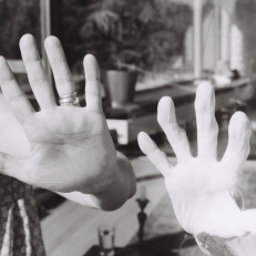Schwarz-Weiß-Fotografie von zwei ausgestreckten Händen, die sich durch eine Glasscheibe berühren, symbolisch für Nähe und Trennung.