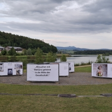 Open-Air-Ausstellung „Demenz neu sehen“ am Seeufer mit Blick auf die Berge, zeigt berührende Motive zum Leben mit Demenz mitten in der Natur.