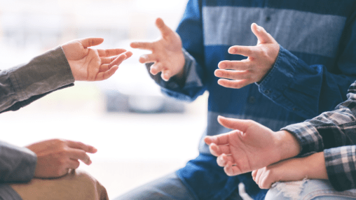 Ausschnitt einer Gesprächssituation in einer Gruppe – mehrere Menschen diskutieren gestikulierend im Kreis, symbolisch für Austausch, Mediation oder Angehörigengespräch.
