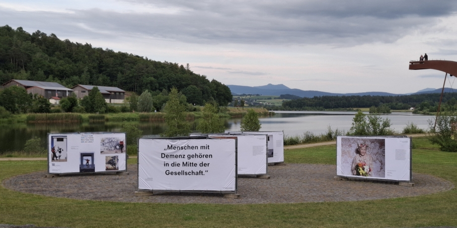 Open-Air-Ausstellung „Demenz neu sehen“ am Seeufer mit Blick auf die Berge, zeigt berührende Motive zum Leben mit Demenz mitten in der Natur.