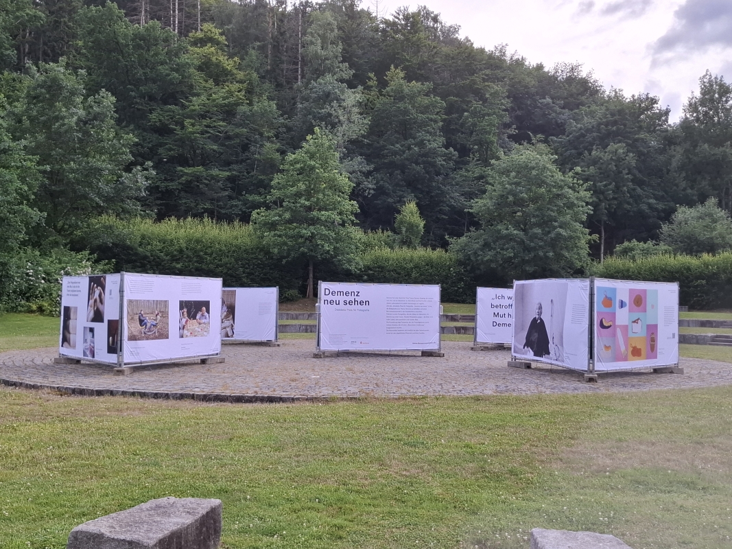 Open-Air-Ausstellung „Demenz neu sehen“ von Desideria Care am Drachensee in Furth im Wald, mit großformatigen Fotobannern inmitten grüner Natur.