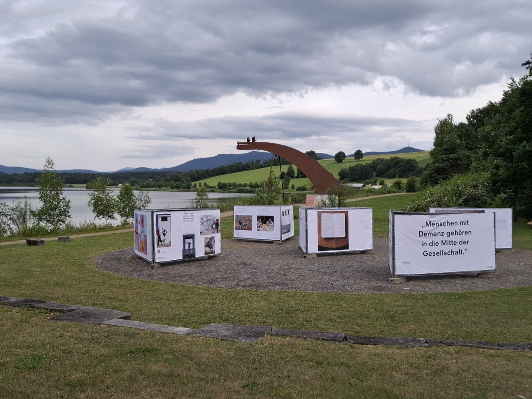 Großformatige Banner der Ausstellung „Demenz neu sehen“ mit emotionalen Fotografien und Zitaten, idyllisch platziert am Drachensee bei Furth im Wald.