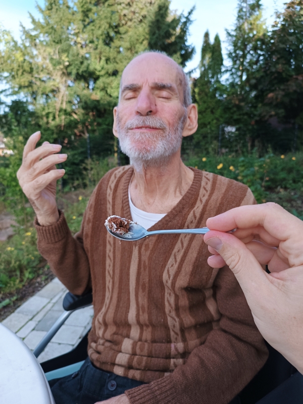 Älterer Mann sitzt im Rollstuhl im Freien, schließt genüsslich die Augen, während ihm jemand mit einem Löffel Essen anbietet.