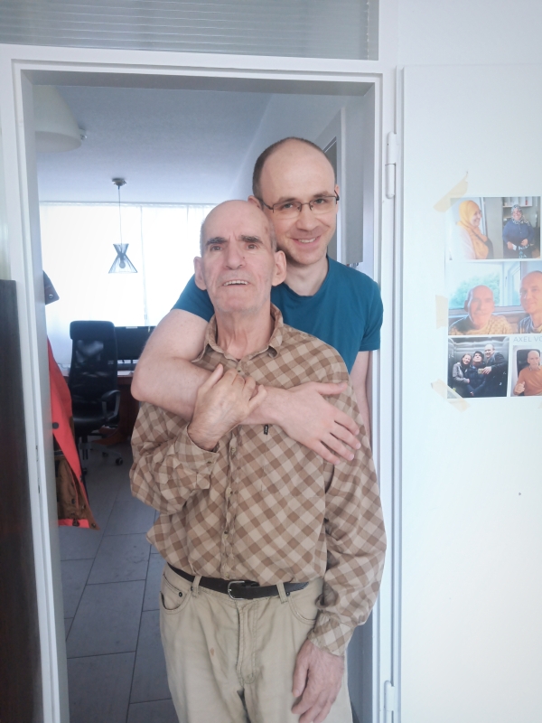Ein jüngerer Mann umarmt von hinten einen älteren Mann, während beide in einem Türrahmen stehen; im Hintergrund ist ein heller Wohnraum zu sehen.