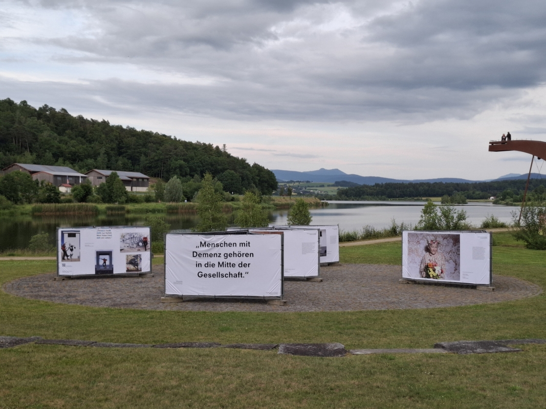 Open-Air-Ausstellung „Demenz neu sehen“ am Seeufer mit Blick auf die Berge, zeigt berührende Motive zum Leben mit Demenz mitten in der Natur.