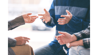 Ausschnitt einer Gesprächssituation in einer Gruppe – mehrere Menschen diskutieren gestikulierend im Kreis, symbolisch für Austausch, Mediation oder Angehörigengespräch.