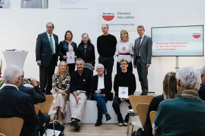 Gruppenfoto von neun Personen auf einer Bühne, eine Person hält eine Urkunde, im Hintergrund das rote Desideria-Logo mit dem Schriftzug 'Demenz neu sehen' und ein Präsentationsbildschirm.