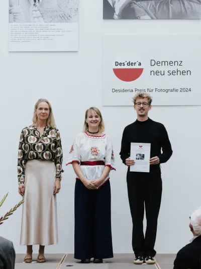 Drei Personen stehen auf einer Bühne, der Mann rechts hält eine Urkunde, im Hintergrund das rote Desideria-Logo mit dem Schriftzug 'Demenz neu sehen'.