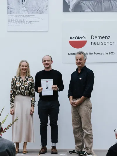 Drei Personen stehen auf einer Bühne, der Mann in der Mitte hält eine Urkunde, im Hintergrund das rote Desideria-Logo mit dem Schriftzug 'Demenz neu sehen'.