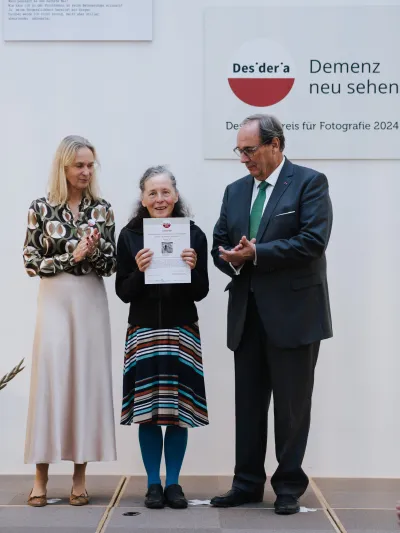 Drei Personen stehen auf einer Bühne, die Frau in der Mitte hält eine Urkunde, links und rechts daneben eine Frau und ein Mann applaudieren, im Hintergrund das rote Desideria-Logo mit dem Schriftzug 'Demenz neu sehen'.