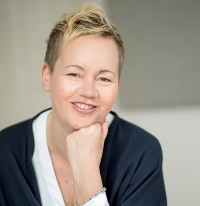 Porträt einer lächelnden Frau mit kurzem blondem Haar, die den Kopf auf ihre Hand stützt, vor neutralem Hintergrund.