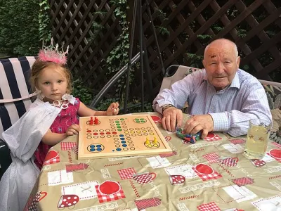 Kleines Mädchen mit Krone und älterer Mann spielen gemeinsam ein Brettspiel im Garten – generationenverbindender Moment zwischen Kind und Großvater, Symbol für Lebensfreude und familiäre Nähe bei Demenz.