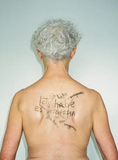 Rückenansicht einer älteren Frau mit weißem Haar und Schriftzug auf der Haut – Ausdruck von Identität, Verletzlichkeit und Demenz