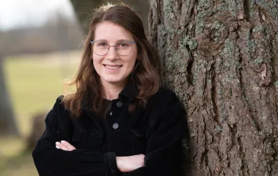 Junge Frau mit Brille lehnt lächelnd an einem Baumstamm in naturnaher Umgebung – freundliches Portraitfoto im Freien, ideal für Team- oder Expertenvorstellungen.