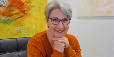 Porträt einer lächelnden Frau mit grauen Haaren und Brille im orangefarbenen Pullover – Expertin im Bereich Demenz und Angehörigenarbeit