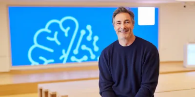 Moderator Marco Schreyl sitzt lächelnd in einem modernen Saal vor einer blauen Leinwand mit stilisierter Gehirn-Grafik. Das Bild entstand anlässlich des DZNE-Podcasts ‚90 Milliarden‘.