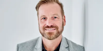 Portrait eines lächelnden Mannes im grauen Sakko vor hellem Hintergrund – professionelles Profilbild vor weißer Wand.