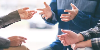 Ausschnitt einer Gesprächssituation in einer Gruppe – mehrere Menschen diskutieren gestikulierend im Kreis, symbolisch für Austausch, Mediation oder Angehörigengespräch.