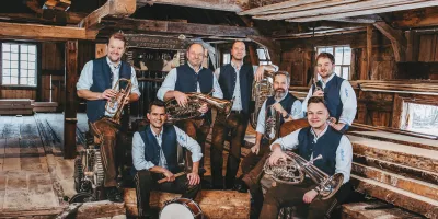 Traditionelle Blasmusikgruppe mit sieben Musikern in Tracht, umgeben von rustikaler Holzhütte – Gruppenbild mit Instrumenten wie Trompete, Tuba, Posaune, Akkordeon und Schlagzeug.