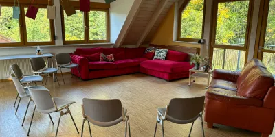 Heller Gruppenraum mit roten Sofas, bequemen Stühlen und großen Fenstern mit Blick ins Grüne, eingerichtet für Gesprächsrunden oder Workshops.