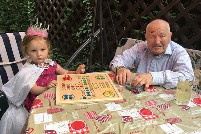 Kleines Mädchen mit Krone und älterer Mann spielen gemeinsam ein Brettspiel im Garten – generationenverbindender Moment zwischen Kind und Großvater, Symbol für Lebensfreude und familiäre Nähe bei Demenz.