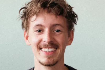 Porträt eines lächelnden jungen Mannes mit lockigem Haar und Bart, der ein dunkles T-Shirt trägt, vor hellem Hintergrund.