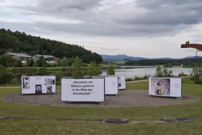 Open-Air-Ausstellung „Demenz neu sehen“ am Seeufer mit Blick auf die Berge, zeigt berührende Motive zum Leben mit Demenz mitten in der Natur.