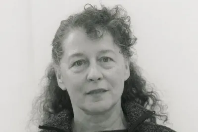 Schwarz-Weiß-Porträt einer Frau mit lockigem, schulterlangem Haar, die direkt in die Kamera blickt, vor hellem Hintergrund.