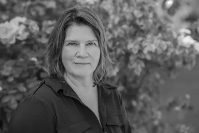 Schwarz-Weiß-Porträt einer Frau mit schulterlangem Haar vor blühenden Sträuchern im Hintergrund.