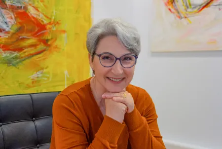 Porträt einer lächelnden Frau mit grauen Haaren und Brille im orangefarbenen Pullover – Expertin im Bereich Demenz und Angehörigenarbeit