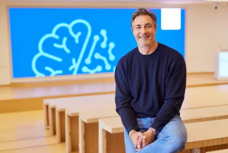 Moderator Marco Schreyl sitzt lächelnd in einem modernen Saal vor einer blauen Leinwand mit stilisierter Gehirn-Grafik. Das Bild entstand anlässlich des DZNE-Podcasts ‚90 Milliarden‘.