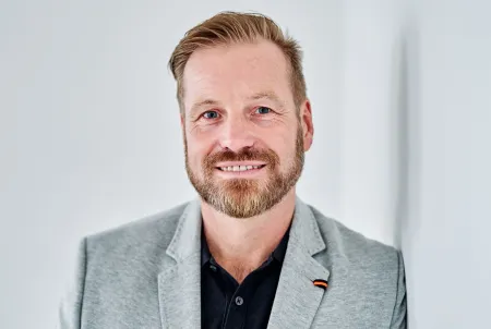 Portrait eines lächelnden Mannes im grauen Sakko vor hellem Hintergrund – professionelles Profilbild vor weißer Wand.