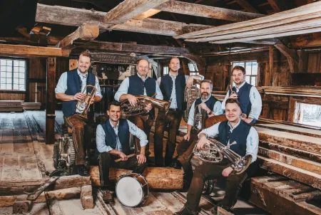 Traditionelle Blasmusikgruppe mit sieben Musikern in Tracht, umgeben von rustikaler Holzhütte – Gruppenbild mit Instrumenten wie Trompete, Tuba, Posaune, Akkordeon und Schlagzeug.