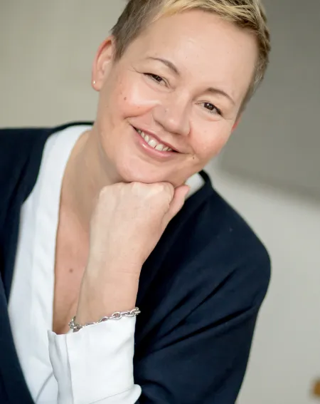 Porträt einer lächelnden Frau mit kurzem blondem Haar, die den Kopf auf ihre Hand stützt, vor neutralem Hintergrund.