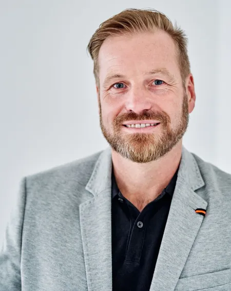 Portrait eines lächelnden Mannes im grauen Sakko vor hellem Hintergrund – professionelles Profilbild vor weißer Wand.