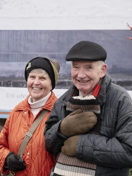 Altes Ehepaar steht Arm in Arm vor einem Plakat.