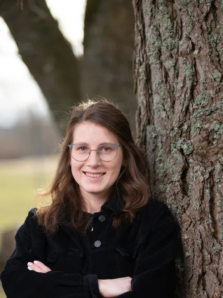 Junge Frau mit Brille lehnt lächelnd an einem Baumstamm in naturnaher Umgebung – freundliches Portraitfoto im Freien, ideal für Team- oder Expertenvorstellungen.